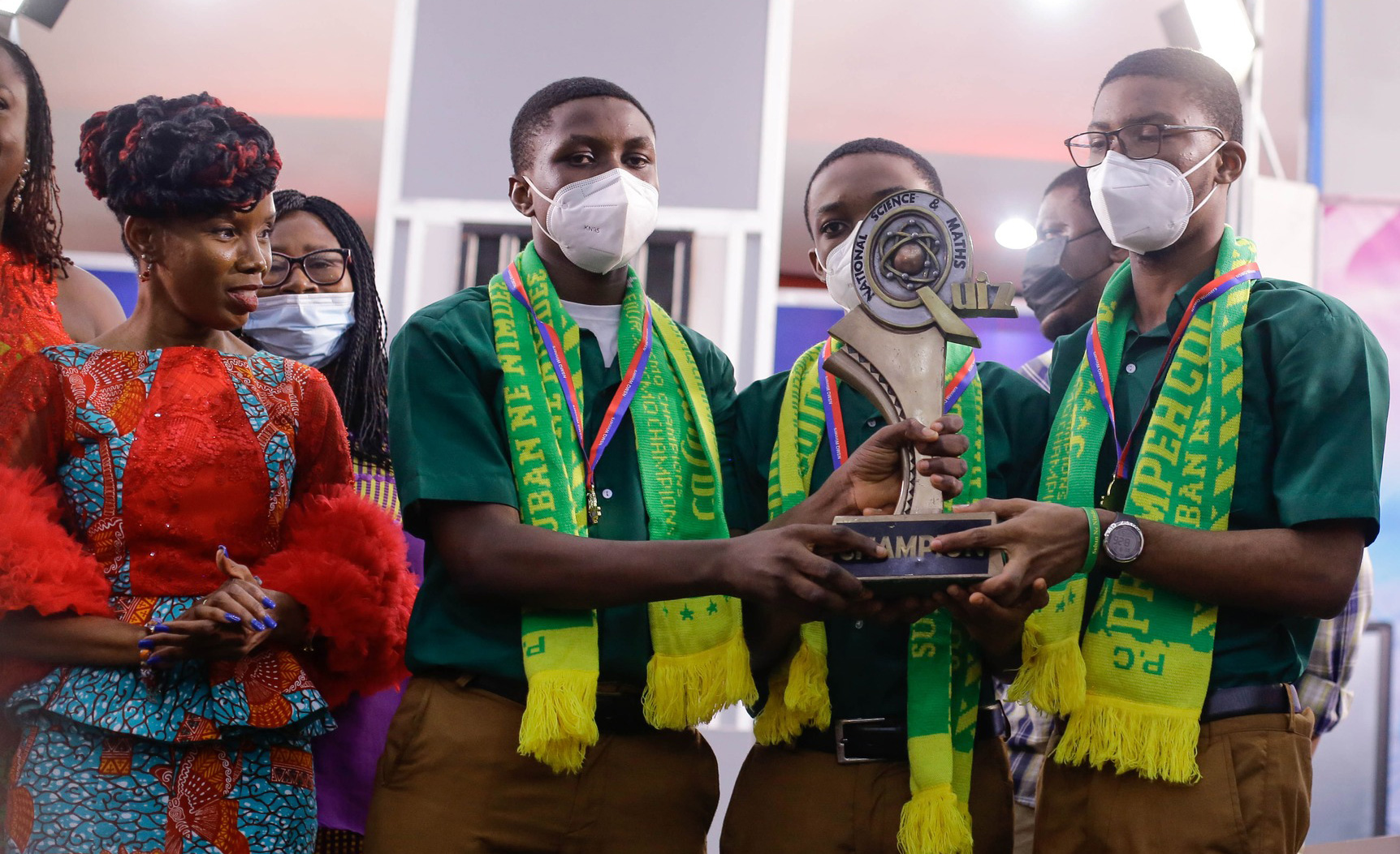 Three Prempeh College students in green shirts and championship sashes hold the National Science and Maths Quiz trophy together on stage.
