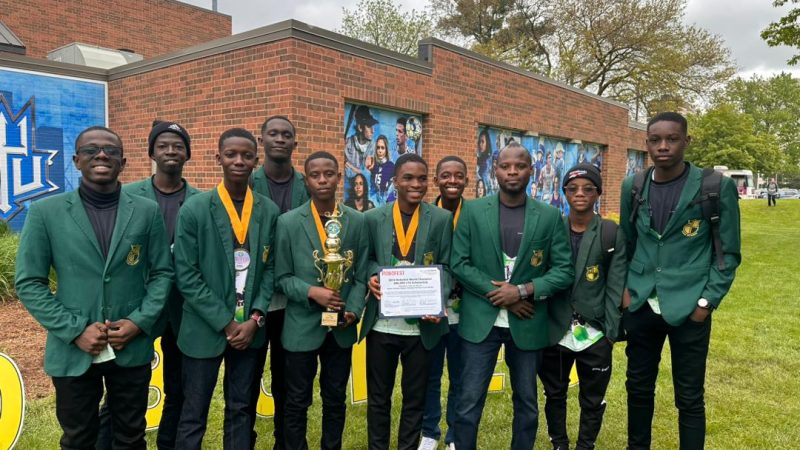 The Prempeh College robotics team in green blazers and medals holds a trophy and a Robofest certificate outside a brick building on a US campus.