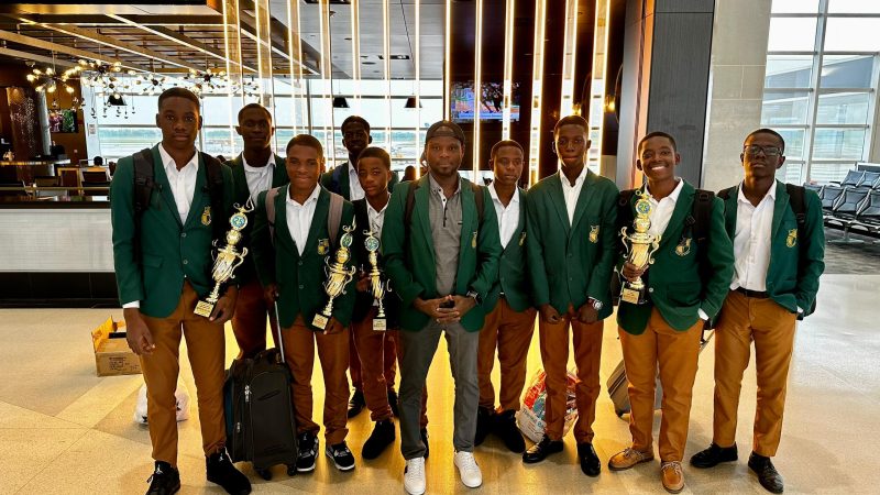 The robotics team in green blazers stands together in an airport terminal, several members holding trophies.